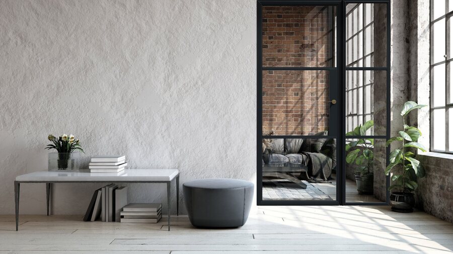 A minimalist interior with a textured white plaster wall, featuring a black-framed glass door that looks into a room with a brick wall and a grey ottoman in the foreground.
