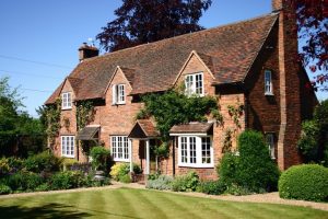 An angled view of a charming, traditional red brick cottage with a multi-gabled tiled roof, white multi-pane windows, climbing ivy, and a well-manicured green lawn out front.